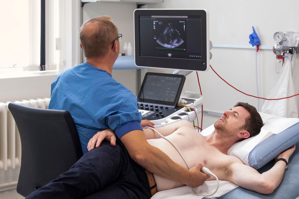 Um profissional de saúde, vestindo uniforme azul, realiza um exame de ecocardiograma em um paciente deitado em uma maca. O médico está de costas, segurando o transdutor de ultrassom contra o peito do homem, enquanto observa atentamente um monitor que exibe imagens em preto e branco do coração em tempo real. O paciente está relaxado, com o braço acima da cabeça, em uma sala de exames clínica bem iluminada e equipada com tecnologia médica avançada.